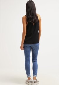 Donna con capelli lunghi e scuri, che indossa una parte superiore nera senza maniche, jeans skinny blu con orli sfilacciati e sneakers argento metallico, in piedi con la schiena rivolta.