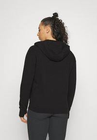 Donna con capelli ricci raccolti a metà in un bun, indossa un hoodie nero e leggins grigi scuri, di spalle su uno sfondo bianco semplice.