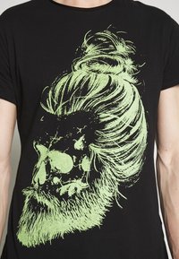 Svart t-shirt med en stor, grön grafisk av en skalle med hår och skägg, som framhäver detaljerade texturer och distinkta linjer.