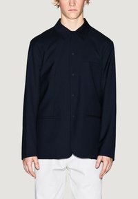 Giacca camicia blu navy con colletto a punta, bottoni frontali e due tasche laterali. Tessuto liscio con una vestibilità su misura e dettagli minimi.