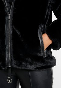 Veste en fausse fourrure noire avec des détails de fermeture éclair argentée et une main dans la poche latérale zippée, associée à un pantalon en cuir noir.
