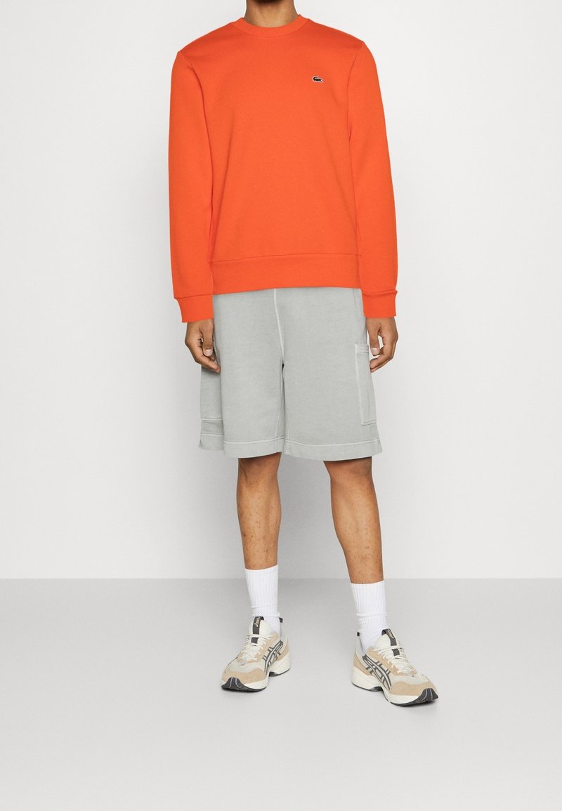 Homme portant un sweat à manches longues orange vif, un short gris clair au genou avec des poches latérales, des chaussettes blanches et des baskets beiges.