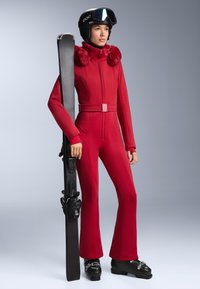 Piros síruházat bordázott textúrával és műszőrme gallérral, deréknál övvel ellátva. Fekete síbakancsok, a bal kézben síléc, fekete sisak viselve.