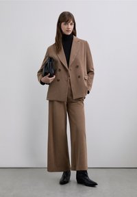 Brauner karierten Anzug mit einem doppelreihigen Blazer und Hosen mit weitem Bein. Kombiniert mit einem schwarzen Rollkragenpullover und einer schwarzen Handtasche. Schwarze Stiefeletten.