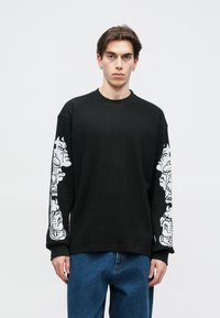 Μαύρο πουλόβερ με μακριά μανίκια από υφή με ραμμένο λαιμό. Διαθέτει λευκά γραφικά σχέδια και στις δύο μανίκες. Φοριέται με μπλε τζιν.