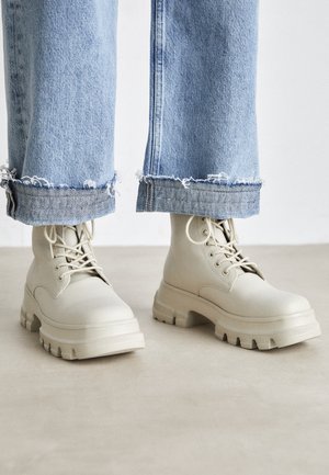 Bottes beige à gros talons avec lacets, portées avec un jean bleu clair retroussé sur un fond neutre.