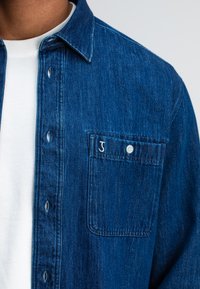 Blauwe denimshirt met een puntkraag, knoopsluiting en een borstzak met een witte geborduurde "J." Daaronder is een witte T-shirt met ronde hals zichtbaar.