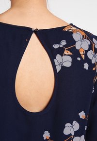 Blouse marine avec un imprimé floral en gris clair et orange, dotée d'un dos à trou de serrure fermé par un petit bouton.