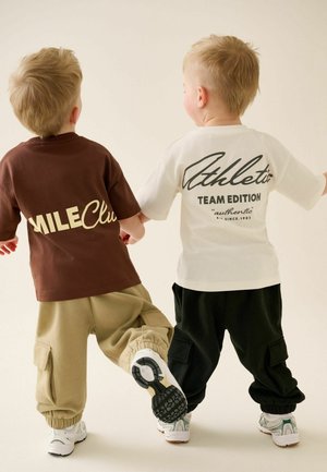 Zwei kleine Kinder mit blondem Haar halten Händchen und tragen legere Hosen sowie T-Shirts mit den Aufdrucken "MILE Club" und "Athletic Team Edition."