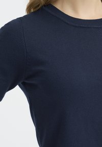 Pull en tricot bleu marine avec un col rond et des manches courtes. Le tissu semble lisse avec une texture et un ajustement uniformes.