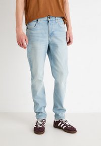 Vaqueros de mezclilla de color azul claro con un diseño de pierna recta, cinco bolsillos y un ligero desgaste. Combinados con zapatillas deportivas burdeos que tienen franjas blancas.