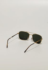 Ochelari de soare pătrați cu ramă din metal auriu, lentile de culoare verde închis și brațe cu accente negre. Prezintă detalii transparente pe ramă.