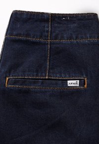 Tela de mezclilla azul oscuro con costuras naranjas que presenta un bolsillo trasero horizontal y una pequeña etiqueta blanca de la marca Levi's.