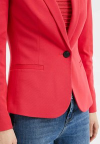 Röd blazer med skräddarsydd design, en svart knapp och två framfickor. Slät tygkvalitet med figurnära siluett. Tillsammans med blå jeans.