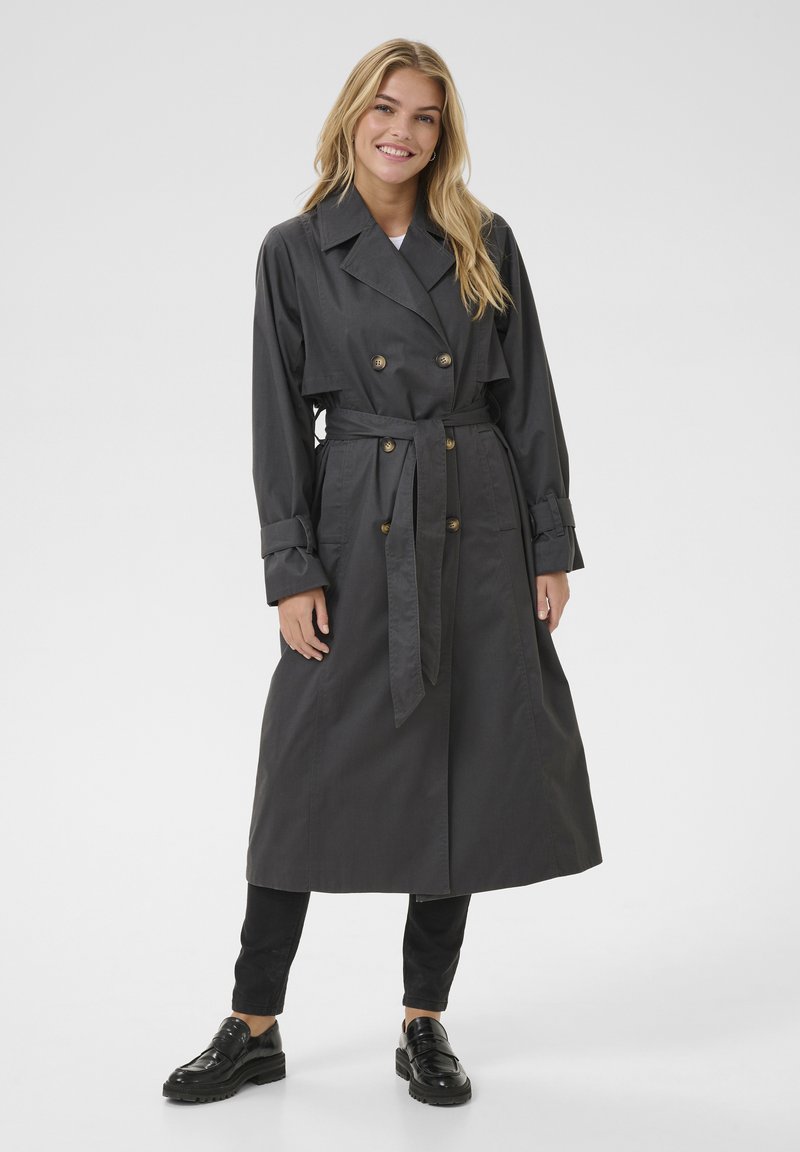 Trench coat grigio scuro con design doppiopetto, vita con cintura e polsini piegati. Modello che indossa pantaloni neri slim e scarpe lucide.