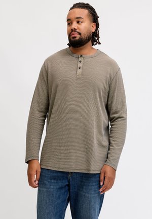 Homme avec des dreadlocks portant un shirt henley gris à manches longues et un jean bleu, debout devant un fond clair uni.