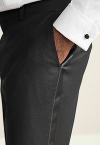 Black tailored trousers featuring a sleek satin side stripe, smooth fabric texture, and a side pocket with a contrasting white shirt cuff.