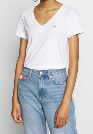 Vrouw draagt een eenvoudige witte V-hals T-shirt, ingestopt in een lichtblauwe high-waisted jeans met een rood-wit-grijs gestreept detail in de zak.