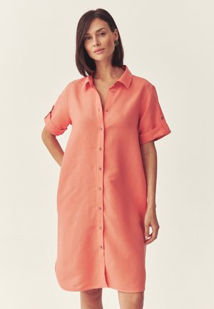Kobieta w koralowej sukience koszulowej z guzikami, z podwiniętymi krótkimi rękawami, stojąca na tle jasnego, jednolitego tła.