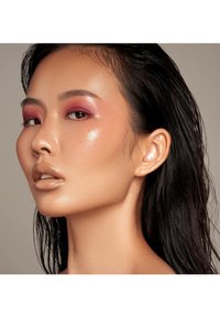 Nærbilde av et ansikt med vått, mørkt hår, glatt hud, dristig rosa øyenskygge, glinsende nudes leppestift og subtile fregner på en nøytral bakgrunn.