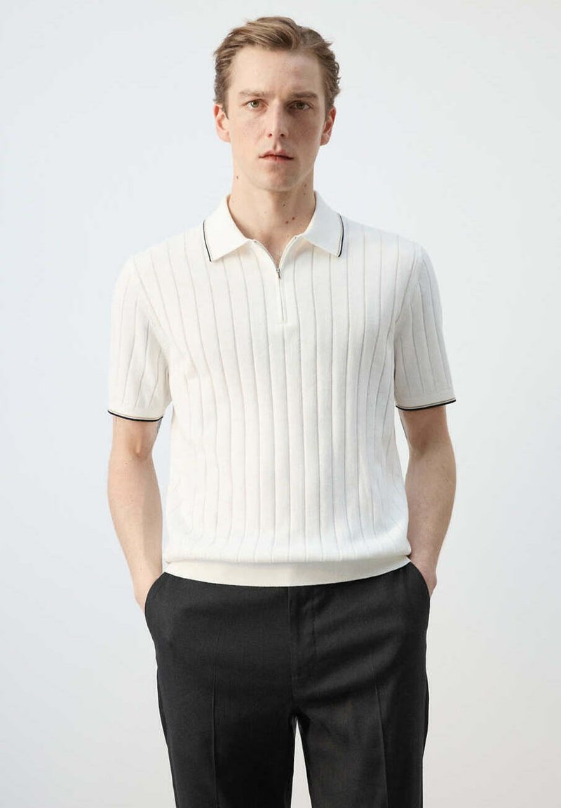 Jeune homme debout les mains dans les poches, portant un polo blanc à manches courtes en maille côtelée et un pantalon noir sur un fond uni.