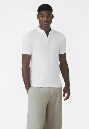 Fehér bordázott galléros póló rövid gallérral és rövid ujjakkal, világosszürke nadrággal párosítva, egyszerű háttér előtt bemutatva.