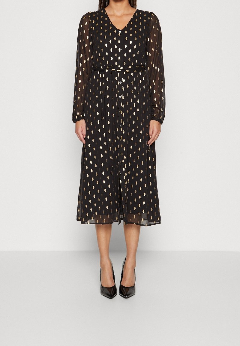 Femme portant une robe midi noire avec des manches longues transparentes et des motifs de feuilles dorées, associée à des escarpins noirs à bouts pointus.