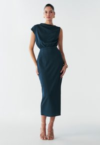 Robe midi bleu sarcelle avec un tissu texturé, un décolleté asymétrique et une taille cintrée. Associée à des sandales dorées à lanières. Silhouette lisse et ajustée.