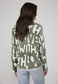 Grüner Strickpullover mit weißem grafischem Text in verschiedenen Größen. Lockere Passform mit Rundhalsausschnitt und langen Ärmeln. Weiche Textur.