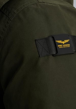 Tissu vert foncé avec une texture lisse. Présente une naperon noir étiqueté "PME LEGEND" avec un texte jaune et un logo d'aile brodé sur l'épaule.