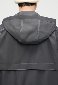 Άτομο που φοράει σκούρο γκρι μπουφάν με κουκούλα και εμφανείς λευκές ραφές, με εκρού πλεκτό καπέλο, εμφανιζόμενο από πίσω σε απλό φόντο.