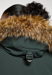 Parka vert foncé avec un col en fausse fourrure épaisse et détachable de couleur marron clair. Dispose d'une boucle noire ajustable au dos du col.