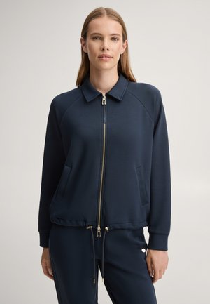 Femme de face, portant une veste zippée bleu marine avec col et un pantalon à cordon assorti, debout devant un fond uni.