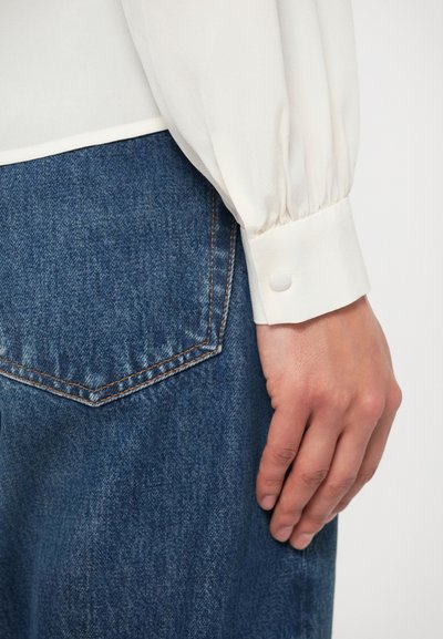 Λευκό, μεταξένιο μπλουζάκι με φουσκωτά μανίκια και μανσέτα με κουμπιά. Συνδυάζεται με μπλε τζιν παντελόνι που έχει ξεχωριστή πίσω τσέπη και πορτοκαλί ραφές.