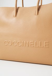 Sac tote Coccinelle en cuir souple couleur tan, avec une surface texturée, un logo en relief et des poignées lisses et structurées.