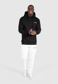 Sort hættetrøje med frontlomme og snoretræk, med hvid branding. Båret sammen med hvide joggere og hvide sneakers. Simpelt, moderne design.