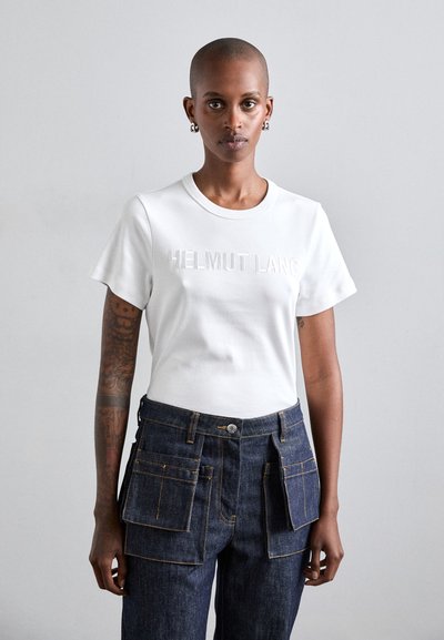 Valkoinen puuvillapaita lyhyillä hihoilla ja kohotettu "HELMUT LANG" -teksti. Yhdistetty tummiin farkkuihin, joissa on useita etutaskuja ja keltainen tikkaus.