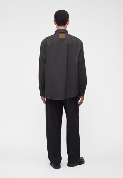 Homme vu de dos portant une chemise ample à manches longues foncée avec une tache brune sur la partie supérieure du dos, un pantalon large noir et des chaussures noires.