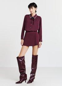 Bordeauxrote Seidenbluse mit perlenbesetztem Kragen, kombiniert mit einem passenden Minirock und glänzenden kniehohen Stiefeln. Durchgehend glatte Textur.