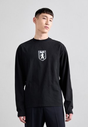 Schwarzes Longsleeve-Shirt aus Baumwolle mit Rundhalsausschnitt. Verfügt über ein zentrales weißes Logo innerhalb eines Wappendesigns.