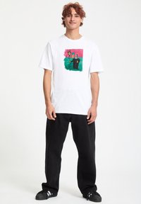 Hvid bomulds T-shirt med et farverigt grafisk design på forsiden, parret med løse sorte jeans og sorte sneakers.