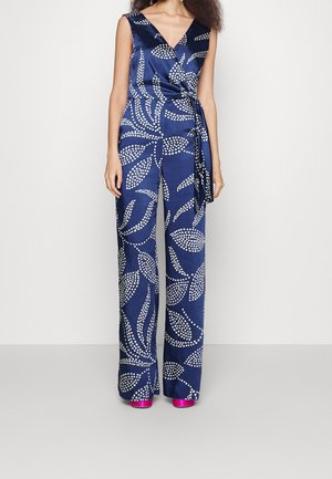 Femme portant une combinaison portefeuille sans manches bleu marine avec des motifs de feuilles blanches à pois et des talons compensés rose vif.