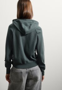 Felpa verde scuro con tasca a canguro e orlo a coste, abbinata a jeans grigi chiari, morbidi e con due tasche posteriori.
