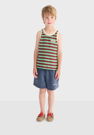 Blond chłopiec stoi, ubrany w zielono-czerwony paskowany tank top, niebieskie szorty i brązowe buty z czerwonymi sznurowadłami na jasnym, jednolitym tle.