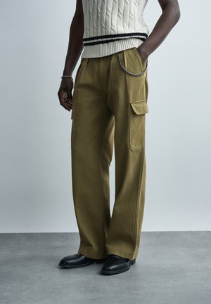 Άνδρας που φοράει χακί παντελόνι cargo με πλαϊνές τσέπες, μαύρα δερμάτινα παπούτσια και λευκό πλεκτό αμάνικο πουλόβερ με μαύρες ρίγες.