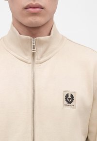 Крупен план на човек, носещ бежово яке с цип и висока яка, с черен нашивка с логото на Belstaff на гърдите.