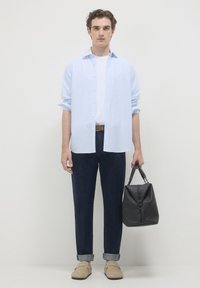 Camicia a righe blu chiaro, t-shirt bianca, pantaloni blu scuro, scarpe beige e una borsa nera testurizzata con manico, posizionata di fronte a uno sfondo semplice.