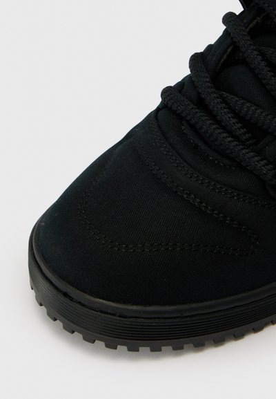 Black athletic shoe with a textured canvas upper, thick black laces, and a rubber outsole featuring a patterned tread design.