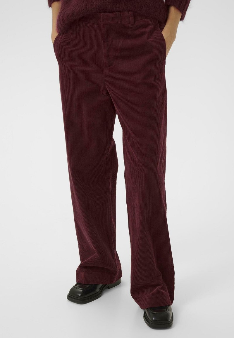 Personne portant un pantalon en velours côtelé rouge foncé et des chaussures noires à bout carré, debout les mains dans les poches devant un fond uni.