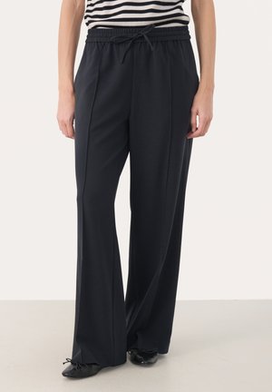 Frau trägt schwarze, weit geschnittene Hose mit elastischem Bund und Kordelzug, kombiniert mit schwarzen Ballerinas und einem gestreiften Oberteil.